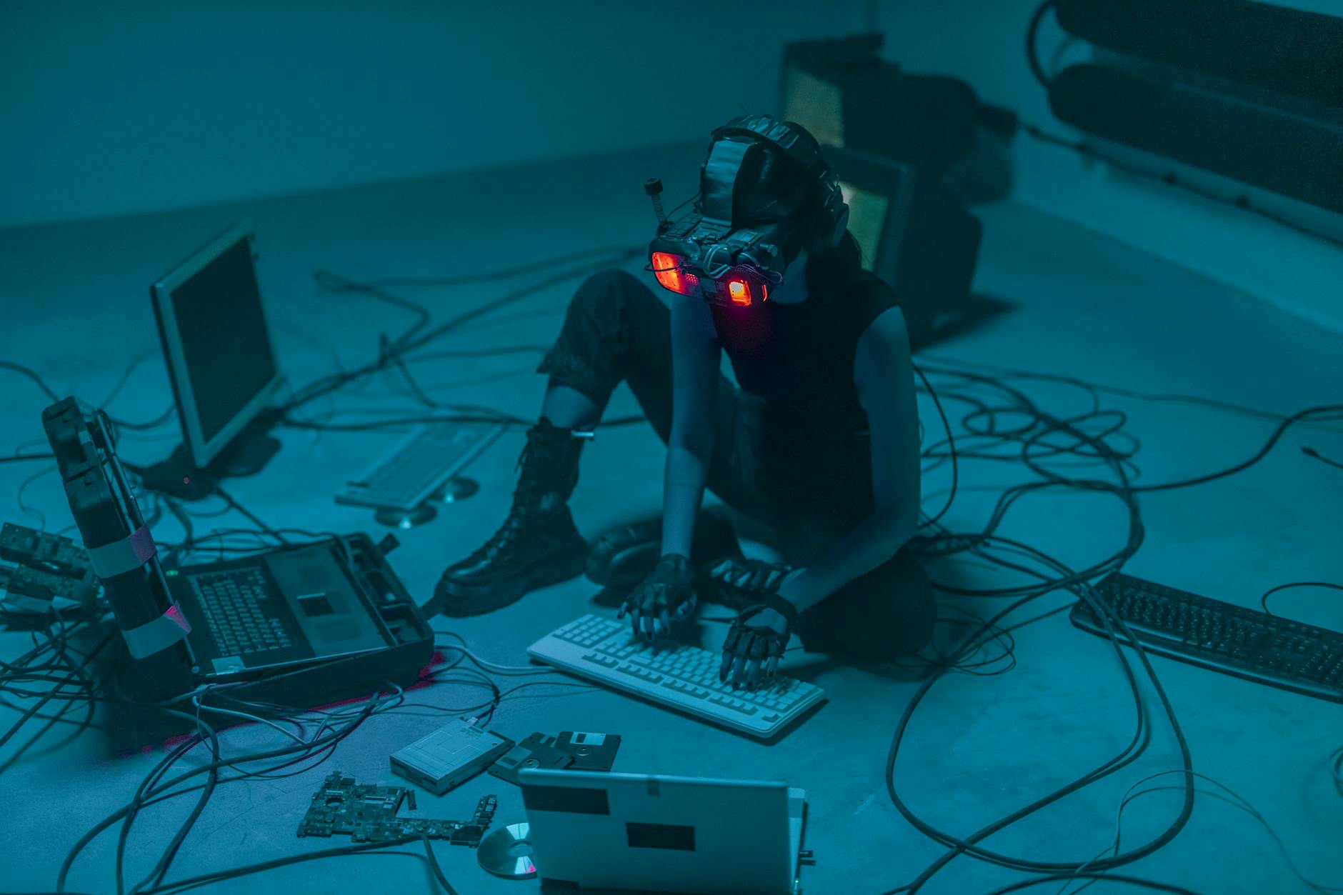 woman sitting on the floor among laptops and tangled cables and wearing goggles